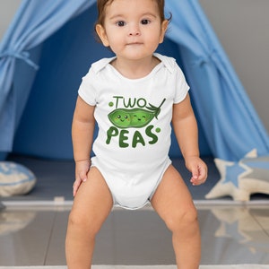 May include: A white baby onesie with a green pea pod graphic and the text "Two Peas". A baby is wearing the onesie and standing on a play mat.