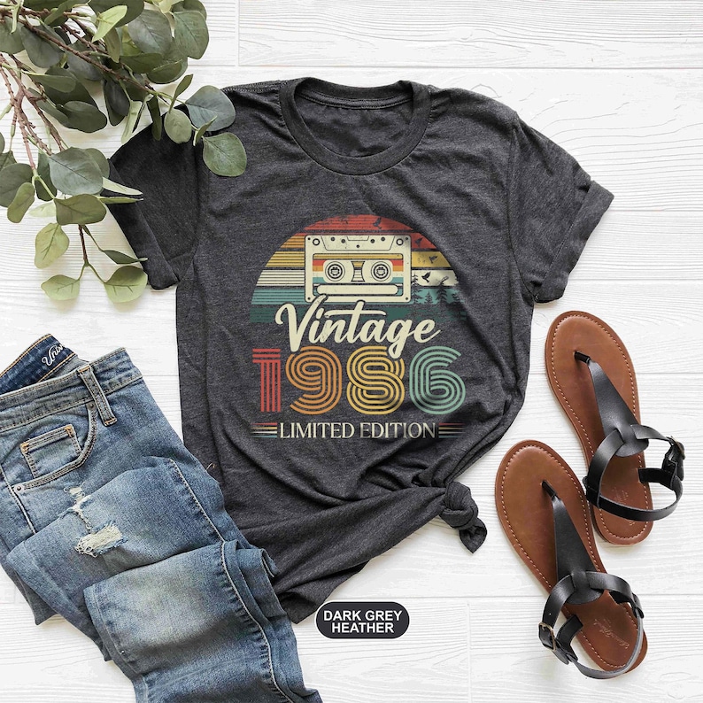 May include: Dark grey heather t-shirt with a retro design featuring a cassette tape and the text "Vintage 1986 Limited Edition." The shirt is paired with blue jeans and brown sandals with black straps.