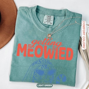 May include: A light green short-sleeve t-shirt featuring the orange text "getting MEOWIED" above a blue line-art illustration of a cat wearing a veil and sunglasses. The shirt has a crew neckline and a Comfort Colors brand label at the inner collar.