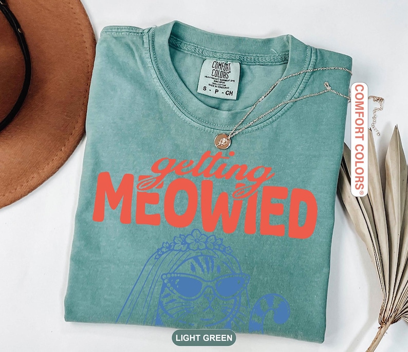 May include: A light green short-sleeve t-shirt featuring the orange text "getting MEOWIED" above a blue line-art illustration of a cat wearing a veil and sunglasses. The shirt has a crew neckline and a Comfort Colors brand label at the inner collar.