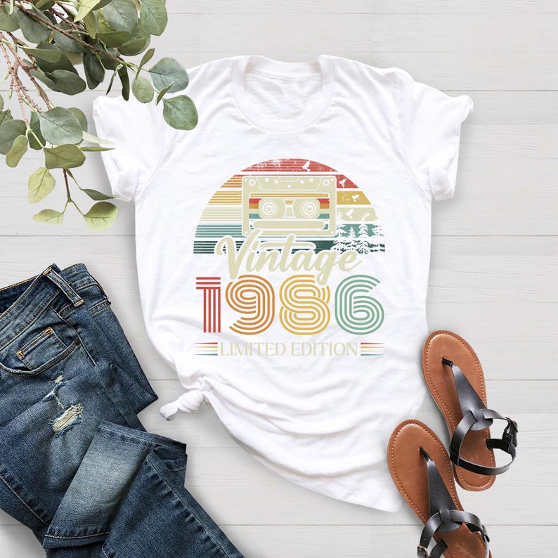 May include: White t-shirt with a retro design featuring a cassette tape, a sunset, and the text "Vintage 1986 Limited Edition" in a colorful, vintage style. The shirt is paired with blue jeans and brown sandals.