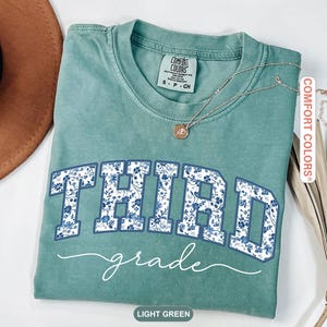 K&ouml;nnte beinhalten: Ein hellgr&uuml;nes T-Shirt mit den Worten "THIRD grade" in einem blauen Blumenmuster. Das Shirt hat einen Rundhalsausschnitt und kurze &Auml;rmel. Eine goldene Halskette mit Anh&auml;nger befindet sich auf dem Shirt.