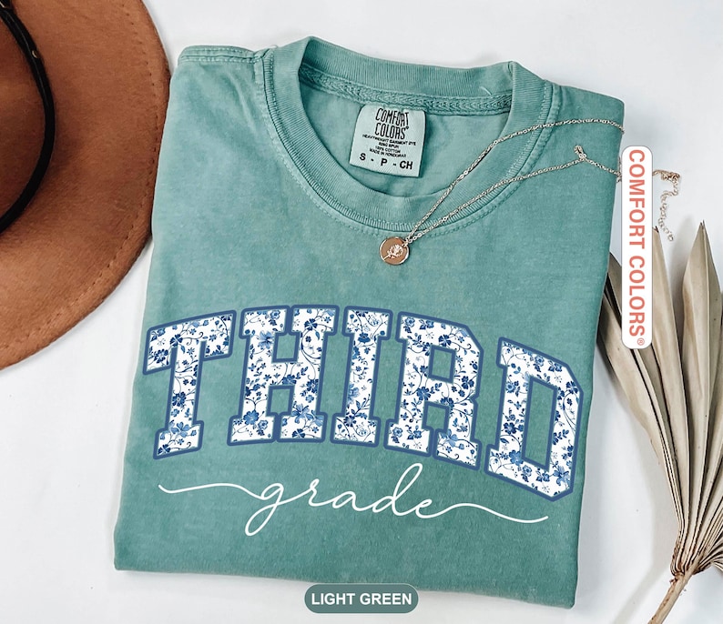 K&ouml;nnte beinhalten: Ein hellgr&uuml;nes T-Shirt mit den Worten "THIRD grade" in einem blauen Blumenmuster. Das Shirt hat einen Rundhalsausschnitt und kurze &Auml;rmel. Eine goldene Halskette mit Anh&auml;nger befindet sich auf dem Shirt.