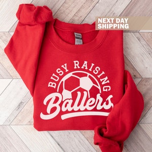 May include: Red sweatshirt with a white graphic that says "Busy Raising Ballers" with a soccer ball in the center.