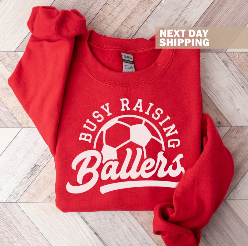 May include: Red sweatshirt with a white graphic that says "Busy Raising Ballers" with a soccer ball in the center.