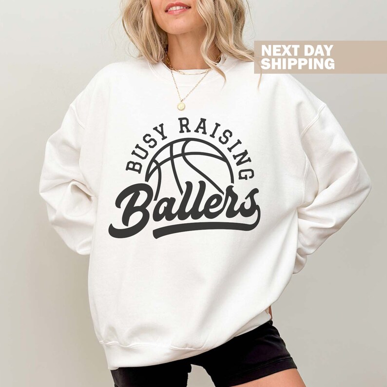 May include: White sweatshirt with black text that reads "Busy Raising Ballers" with a basketball graphic.