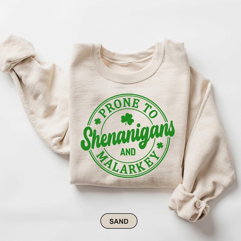 Puede incluir: Una sudadera beige con un gr&aacute;fico circular verde que dice "Prone to Shenanigans and Malarkey" con un dise&ntilde;o de tr&eacute;bol.