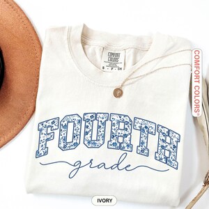 May include: Ivory-colored Comfort Colors t-shirt with "FOURTH" in blue floral print block letters and "grade" in a cursive font. A gold necklace with a pendant is draped on the shirt. The shirt is folded and placed on a white surface.