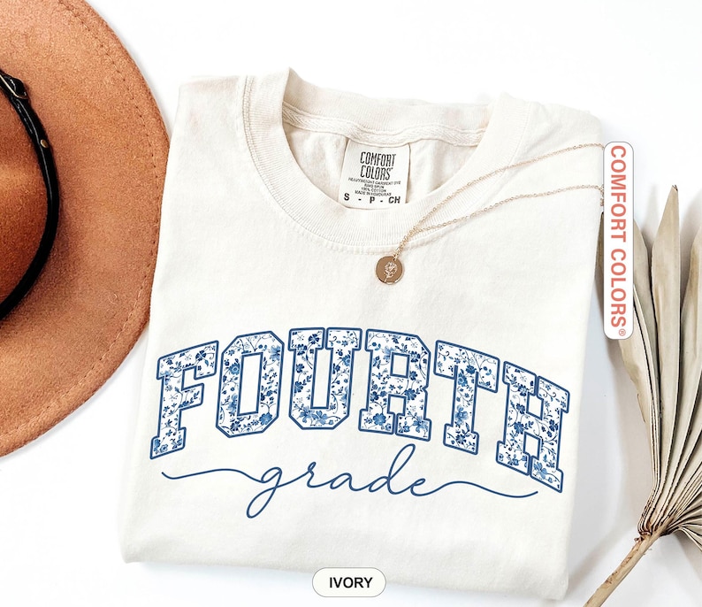 May include: Ivory-colored Comfort Colors t-shirt with "FOURTH" in blue floral print block letters and "grade" in a cursive font. A gold necklace with a pendant is draped on the shirt. The shirt is folded and placed on a white surface.