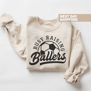 May include: A beige sweatshirt with the text "Busy Raising Ballers" in black lettering. A black and white soccer ball is in the center of the text.