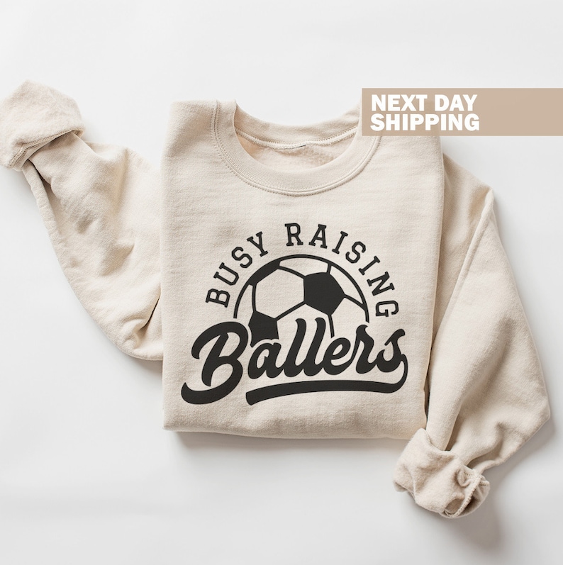 May include: A beige sweatshirt with the text "Busy Raising Ballers" in black lettering. A black and white soccer ball is in the center of the text.