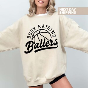 May include: A beige sweatshirt with a black basketball graphic and the text "Busy Raising Ballers".