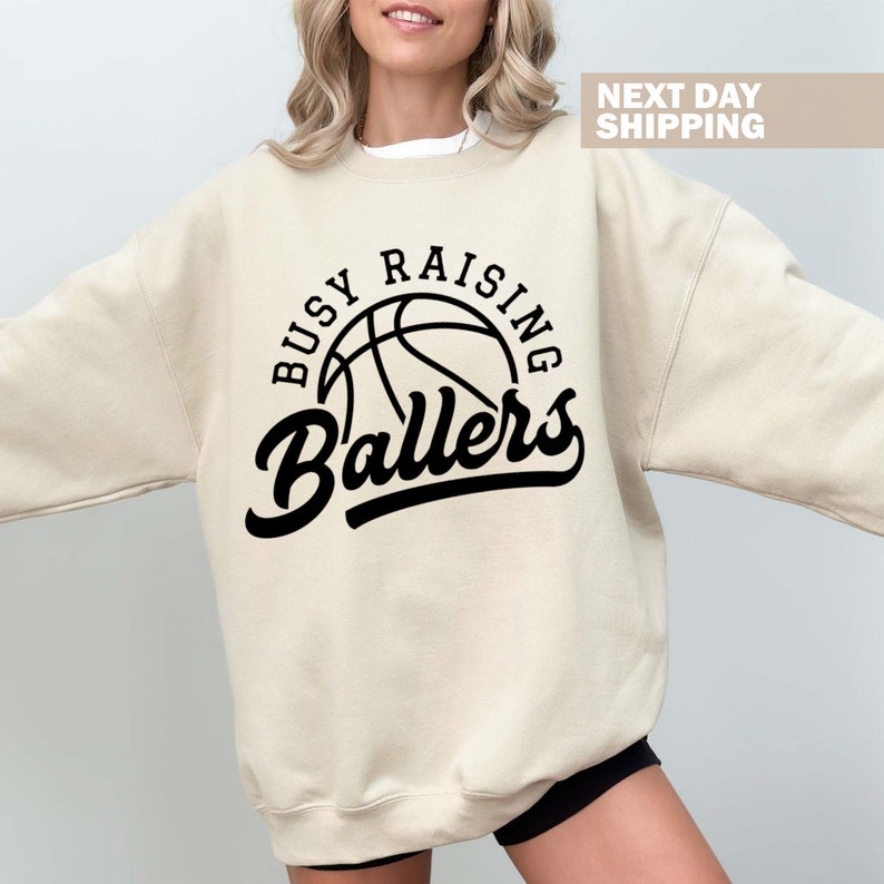 May include: A beige sweatshirt with a black basketball graphic and the text "Busy Raising Ballers".