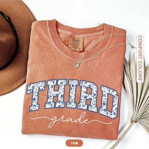 K&ouml;nnte beinhalten: Ein korallenfarbenes Sweatshirt mit Rundhalsausschnitt und den Worten "THIRD grade" in einem blauen Blumenmuster. Eine goldene Halskette und ein brauner Hut sind sichtbar. Das Sweatshirt ist von Comfort Colors.