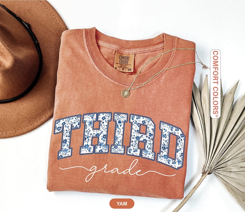 K&ouml;nnte beinhalten: Ein korallenfarbenes Sweatshirt mit Rundhalsausschnitt und den Worten "THIRD grade" in einem blauen Blumenmuster. Eine goldene Halskette und ein brauner Hut sind sichtbar. Das Sweatshirt ist von Comfort Colors.