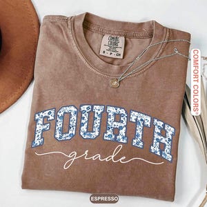 May include: Espresso-colored t-shirt with the words "FOURTH grade" in blue floral print. The shirt has a crew neck and short sleeves. A silver necklace with a pendant is draped on the shirt.