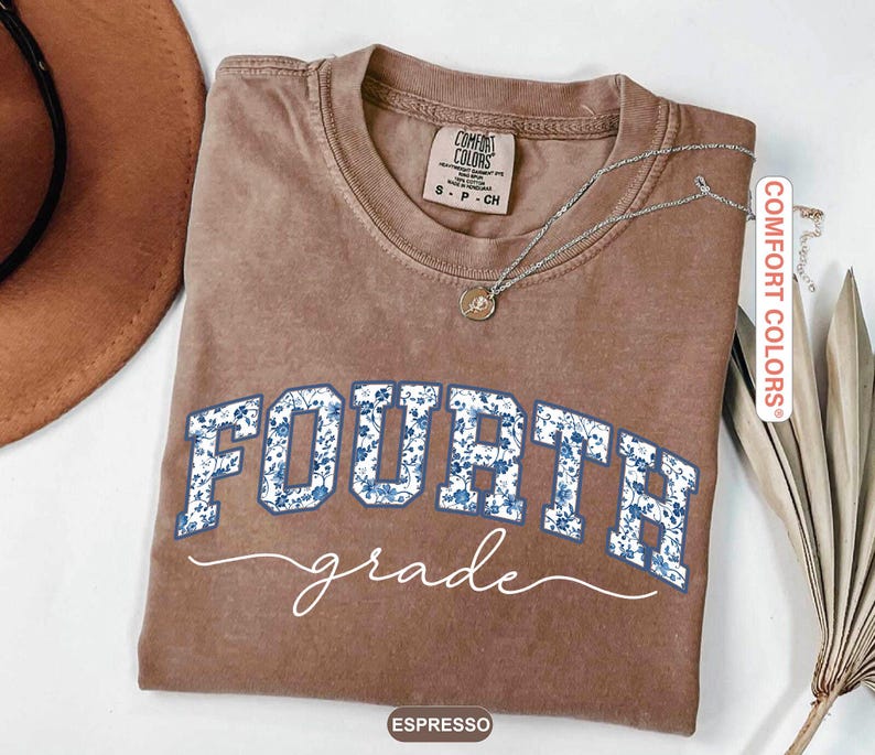May include: Espresso-colored t-shirt with the words "FOURTH grade" in blue floral print. The shirt has a crew neck and short sleeves. A silver necklace with a pendant is draped on the shirt.
