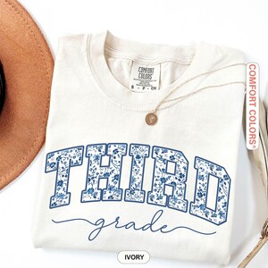 K&ouml;nnte beinhalten: Elfenbeinfarbenes T-Shirt mit dem Schriftzug "THIRD grade" in blauem Blumenmuster. Das Shirt hat einen Rundhalsausschnitt und kurze &Auml;rmel. Eine goldene Halskette mit Anh&auml;nger liegt auf dem Shirt. Die Marke "Comfort Colors" ist sichtbar.