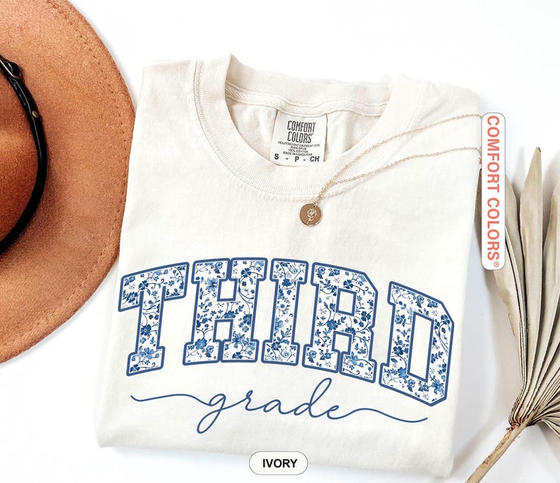 K&ouml;nnte beinhalten: Elfenbeinfarbenes T-Shirt mit dem Schriftzug "THIRD grade" in blauem Blumenmuster. Das Shirt hat einen Rundhalsausschnitt und kurze &Auml;rmel. Eine goldene Halskette mit Anh&auml;nger liegt auf dem Shirt. Die Marke "Comfort Colors" ist sichtbar.