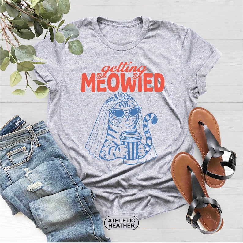 May include: An athletic heather gray t-shirt featuring a blue graphic of a cat wearing a bridal veil and sunglasses while holding a coffee cup. The text "getting MEOWIED" is printed in red and orange above the cat. A small label reads "ATHLETIC HEATHER".