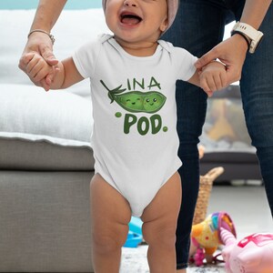May include: A white baby onesie with a green pea pod design. The pea pod has two smiling faces inside and the text "INA POD" is written above it.