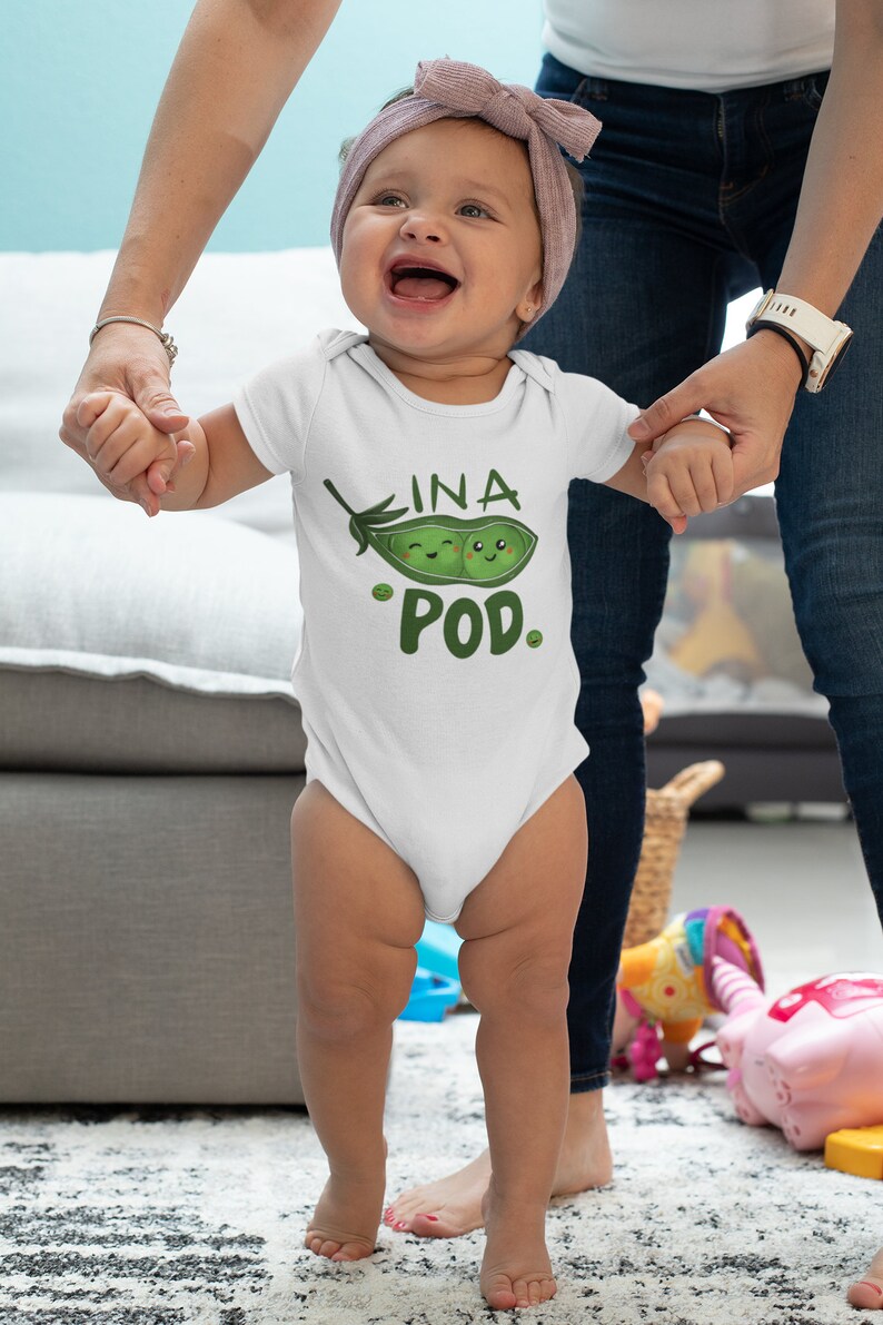 May include: A white baby onesie with a green pea pod design. The pea pod has two smiling faces inside and the text "INA POD" is written above it.