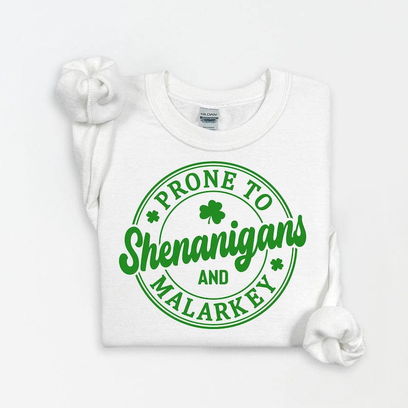 Puede incluir: Camiseta blanca con un gr&aacute;fico circular verde que dice "Prone to Shenanigans and Malarkey" con dise&ntilde;os de tr&eacute;boles.