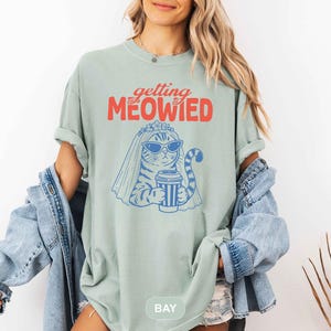 May include: A sage green t-shirt featuring a graphic of a cat wearing sunglasses and a bridal veil while holding a coffee cup. The text above the cat reads "getting MEOWIED" in red and orange lettering. The cat illustration is rendered in blue.