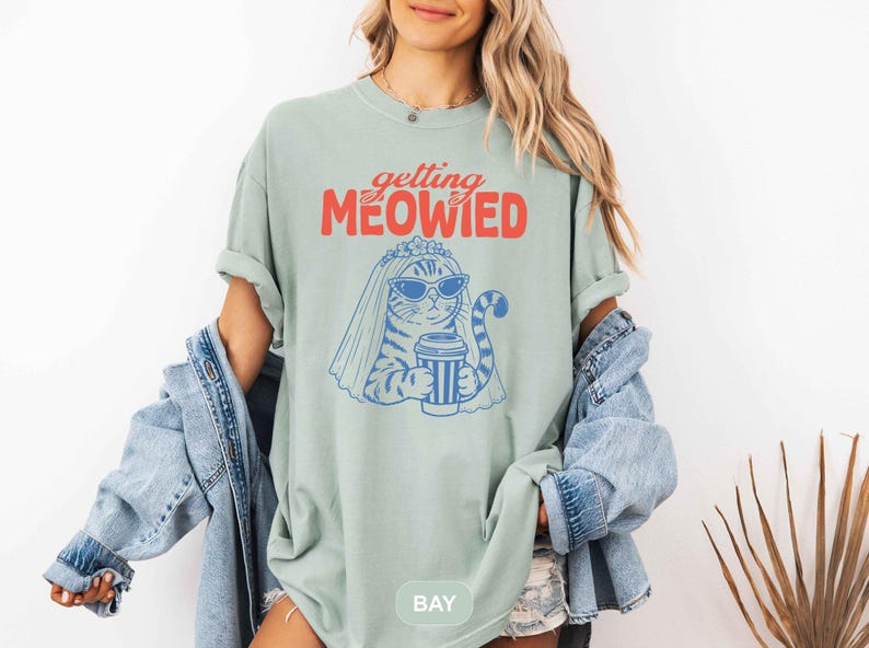May include: A sage green t-shirt featuring a graphic of a cat wearing sunglasses and a bridal veil while holding a coffee cup. The text above the cat reads "getting MEOWIED" in red and orange lettering. The cat illustration is rendered in blue.