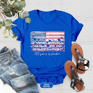 May include: A royal blue t-shirt featuring a watercolor American flag design with floral accents in red, white, and blue. The text "250 years of freedom" is printed below the flag. The shirt is paired with blue jeans and brown sandals.