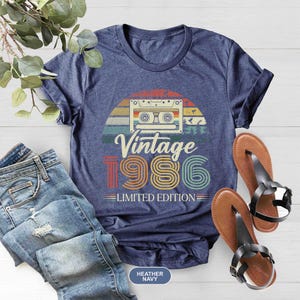 May include: Heather navy blue t-shirt with a retro design featuring a cassette tape and the text "Vintage 1986 Limited Edition." The shirt is paired with blue jeans and brown sandals.
