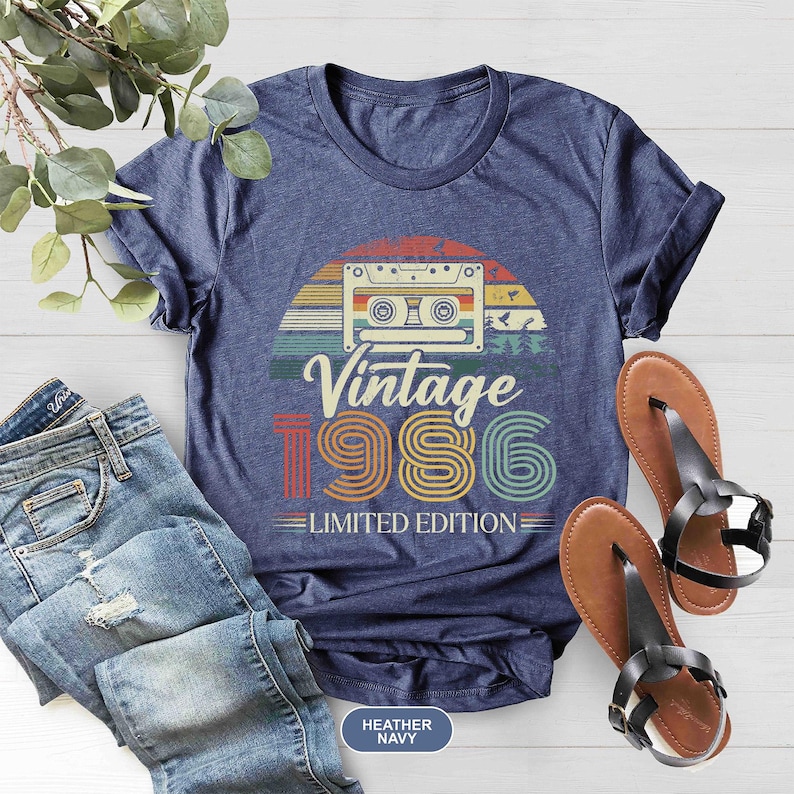 May include: Heather navy blue t-shirt with a retro design featuring a cassette tape and the text "Vintage 1986 Limited Edition." The shirt is paired with blue jeans and brown sandals.