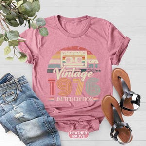 May include: A heather mauve t-shirt featuring a vintage cassette tape design. The graphic includes the words "Vintage 1976 Limited Edition" in a retro font, with a colourful sunset background. The shirt is paired with blue jeans and brown sandals.