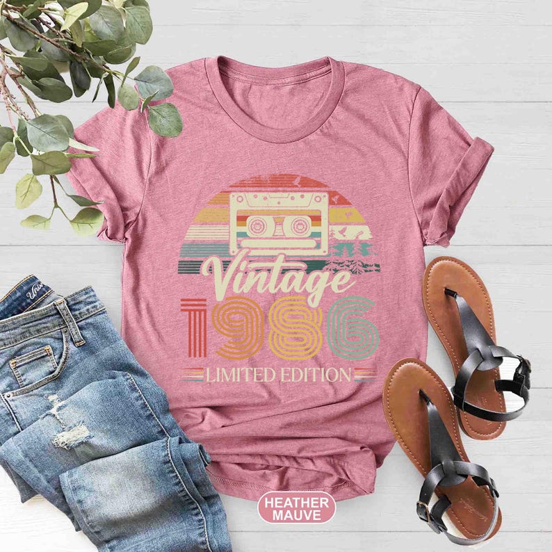 May include: A heather mauve t-shirt with a retro cassette tape design and the text "Vintage 1986 Limited Edition." The shirt is paired with blue jeans and brown sandals with black straps. A sprig of greenery is in the upper left corner.