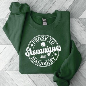 Puede incluir: Una sudadera verde oscuro con un gr&aacute;fico circular blanco que dice "Prone to Shenanigans and Malarkey" con un tr&eacute;bol dentro del c&iacute;rculo.