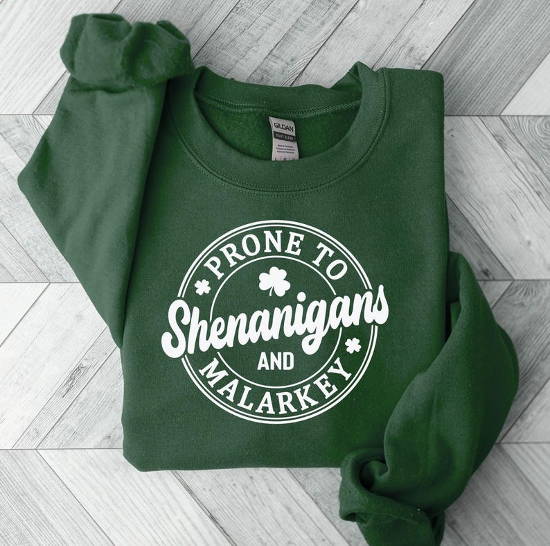 Puede incluir: Una sudadera verde oscuro con un gr&aacute;fico circular blanco que dice "Prone to Shenanigans and Malarkey" con un tr&eacute;bol dentro del c&iacute;rculo.