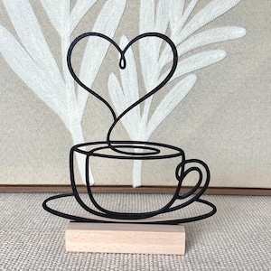 Könnte beinhalten: Schwarze Metallskulptur einer Kaffeetasse mit Untertasse und herzförmigem Dampf. Auf einem kleinen Holzsockel montiert. Der Hintergrund zeigt eine neutrale Leinwand mit weißen blattartigen Formen. Die Skulptur ist ca. 10 cm hoch.