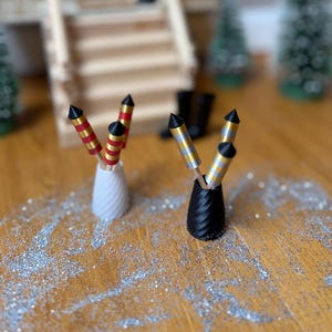 Könnte beinhalten: Zwei Miniatur-Feuerwerksdisplays, eines in einer weißen Vase und das andere in einer schwarzen Vase, stehen auf einer Holzoberfläche, die mit Glitzer bestreut ist. Die Feuerwerke haben rote und goldene Akzente. Ein Miniaturhaus und Weihnachtsbäume sind im Hintergrund.