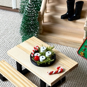 May include: Miniature Christmas scene featuring a wooden picnic table with a black base. A black tray holds four white candles, red berries, and greenery. Candy canes and miniature Christmas trees complete the festive display. Black boots sit on the porch steps.