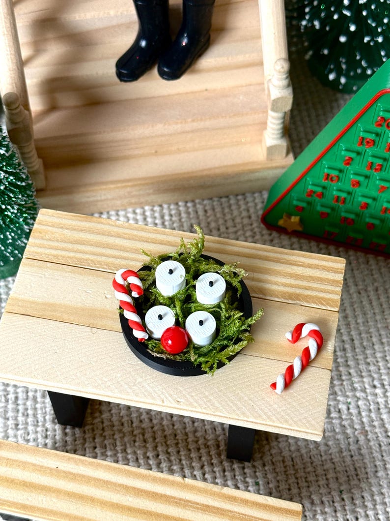 May include: Miniature Christmas scene featuring a wooden picnic table with a black bowl holding white candles and a red ball, surrounded by green moss. Two candy canes and a Christmas tree are also visible.