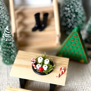 May include: A miniature Christmas scene featuring a small wooden picnic table with a black rimmed plate holding white candles, a red ball, and candy canes. Black boots sit on a wooden staircase in the background, with miniature Christmas trees.