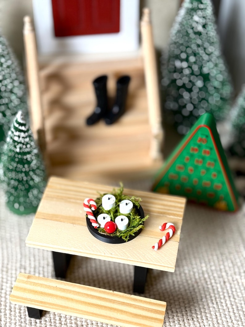 May include: A miniature Christmas scene featuring a small wooden picnic table with a black rimmed plate holding white candles, a red ball, and candy canes. Black boots sit on a wooden staircase in the background, with miniature Christmas trees.