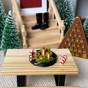 May include: Miniature Christmas scene with a wooden picnic table holding a black tray with gold candles and greenery. Two candy canes and a brown advent calendar are also present. Green Christmas trees and a house with black boots on the steps are in the background.