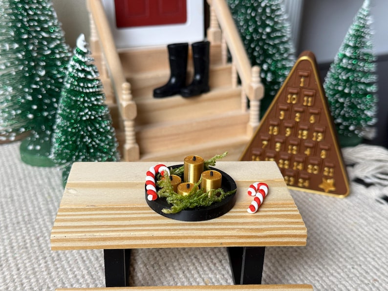 May include: Miniature Christmas scene with a wooden picnic table holding a black tray with gold candles and greenery. Two candy canes and a brown advent calendar are also present. Green Christmas trees and a house with black boots on the steps are in the background.