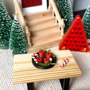 May include: Miniature Christmas scene featuring a wooden table with a black tray holding red candles and greenery. Candy canes are placed on the table. A red triangular advent calendar and miniature Christmas trees are in the background.