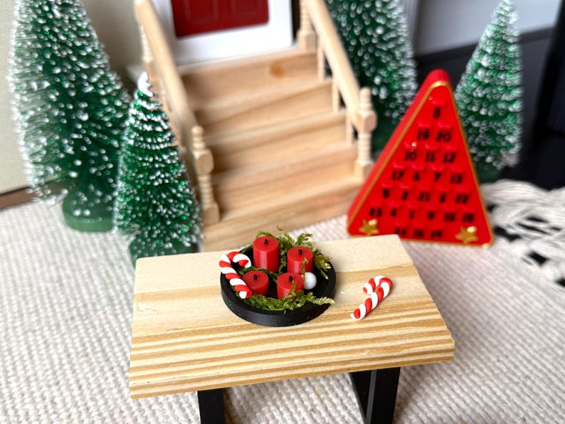 May include: Miniature Christmas scene featuring a wooden table with a black tray holding red candles and greenery. Candy canes are placed on the table. A red triangular advent calendar and miniature Christmas trees are in the background.