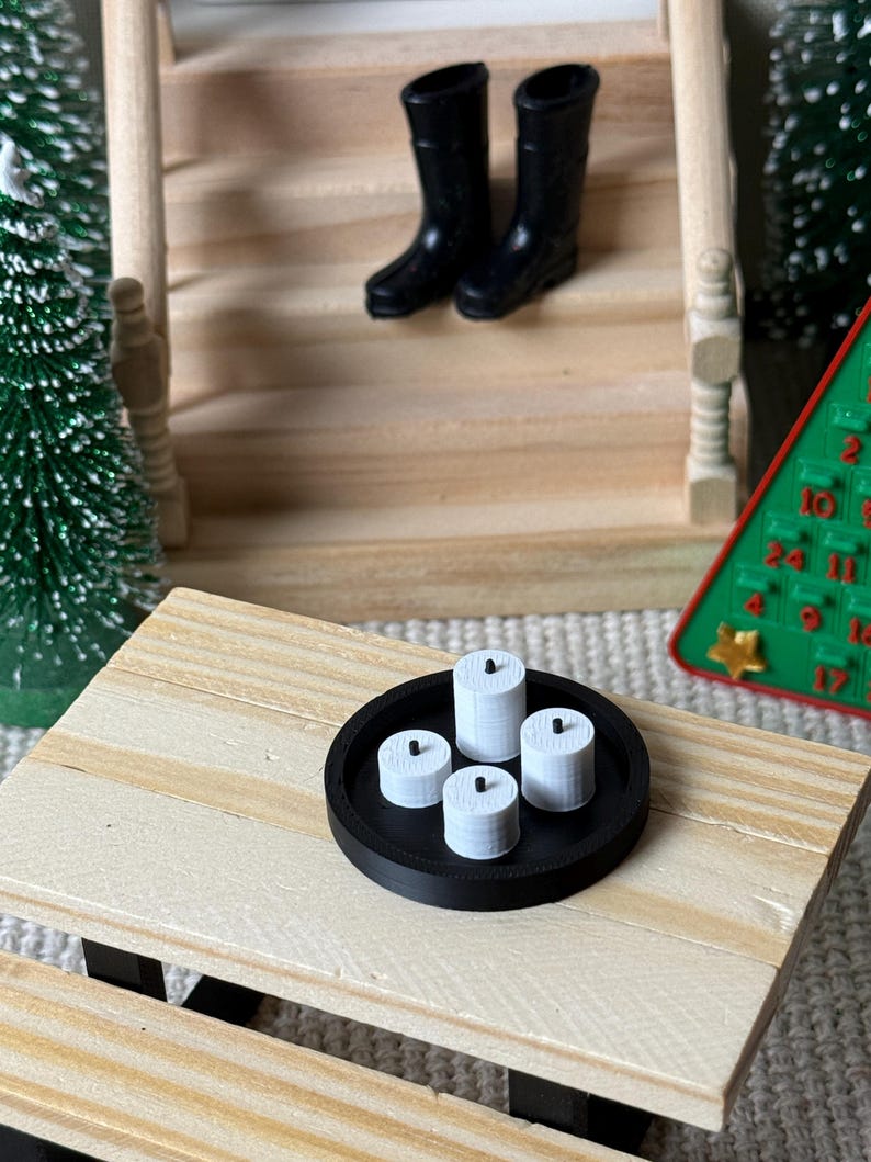 May include: Miniature scene with a black tray holding four white candles. Two black boots are on the wooden stairs in the background. A small Christmas tree and a green advent calendar are also visible.