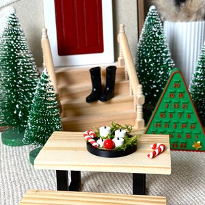 May include: Miniature Christmas scene with a wooden picnic table, black boots, and a red door. The table holds a candle arrangement and candy canes. Green Christmas trees and a green advent calendar complete the festive display.