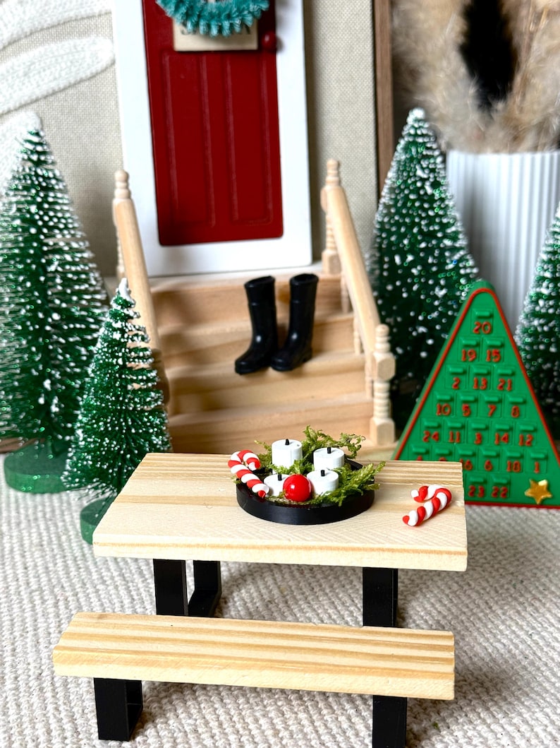 May include: Miniature Christmas scene with a wooden picnic table, black boots, and a red door. The table holds a candle arrangement and candy canes. Green Christmas trees and a green advent calendar complete the festive display.