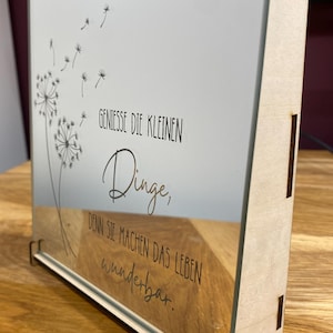 Könnte beinhalten: Ein Holzrahmen mit einem Glasspiegel im Inneren. Der Spiegel hat ein Löwenzahnmuster und den Text "Geniesse die kleinen Dinge, denn sie machen das Leben wunderbar." Der Rahmen steht auf einer Holzoberfläche.